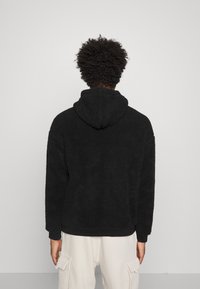 Hanorac din fleece negru, pufos, cu o textură moale, glugă cu șnur și manșete cu nervuri, purtat cu pantaloni cargo de culoare deschisă.