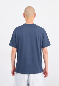 Άτομο με ξυρισμένο κεφάλι, ντυμένο με μπλε μπλούζα και λευκές βερμούδες, στραμμένο προς τα πίσω έναντι ενός απλού λευκού φόντου.