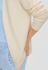 Beige textuur cardigan met geribde manchetten, gedrapeerd ontwerp, gedragen over een lichtblauwe top en gecombineerd met pastelblauwe broek.