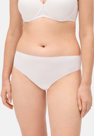 Mujer vistiendo un sujetador de encaje blanco y ropa interior sin costuras a juego, de pie con los brazos relajados contra un fondo claro y liso.