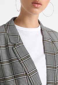 Mustavalkoinen ruudullinen bleiseri, jossa on kultaisia yksityiskohtia, ja joka on päällystetty valkoisella ribbatulla paidalla. Suuret renkaat korvakorut ovat näkyvissä.