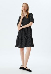 Vestido negro con lunares blancos, mangas cortas, cuello abotonado, falda de volantes, tejido suave; combinado con bailarinas negras.