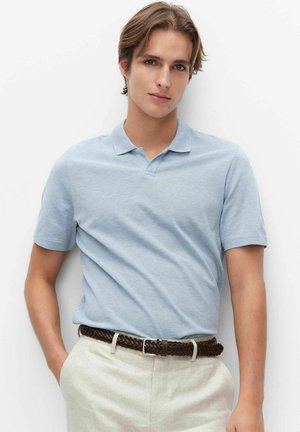 Jonge man draagt een lichtblauw polo met korte mouwen, beige broek en een gevlochten bruine riem, poseert met één hand in zijn zak.