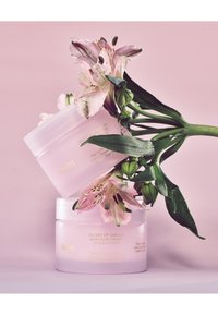 Twee doorschijnende roze potten Huxley huidcrème met gouden letters, genesteld tussen roze bloemen met zichtbare groene bladeren op een roze achtergrond.