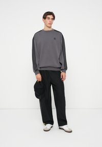 Grå sweatshirt med svarta ränder på ärmarna, som har en liten svart logotyp. Matcher med lösa svarta byxor och vita sneakers, håller en keps.