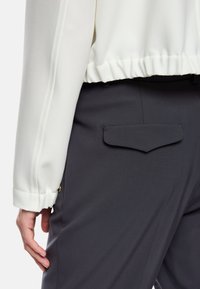 Weiße Bluse mit gerafftem Saum und langen Ärmeln, kombiniert mit dunkelgrauen Hosen, die eine kleine Gesäßtasche und eine glatte Textur aufweisen.