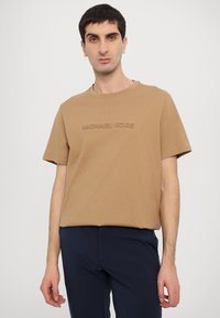 Brown cotton t-shirt featuring a crew neck, short sleeves, and an embossed "MICHAEL KORS" logo on the front. Styled with navy pants.
