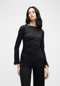 Γυναίκα με μακριά σκούρα μαλλιά, φορώντας μαύρο εφαρμοστό τοπ με διάφανα μακριά μανίκια και μαύρο παντελόνι, στέκεται μπροστά σε ένα απλό φωτεινό φόντο.