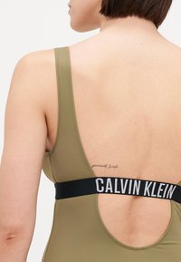 Olíva zöld fürdőruha alacsony hátrészes és széles pántokkal. Fekete elasztikus szalaggal rendelkezik, rajta a "CALVIN KLEIN" logóval. Simább textúra, minimális részletezéssel.
