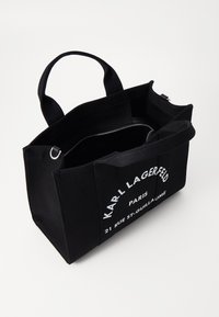 Sort lærredstaske med to øverste håndtag, med hvidt trykt tekst. Indvendigt er der en lynlås lomme. Robust rektangulær form.