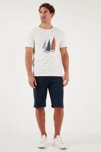 Mees, kes kannab valget T-särki, millel on kolm purjekat graafilist kujutist, tumelisi lühikesi pükse ja valgeid vabaajajalatsid, seisab tavalise heleda tausta ees.