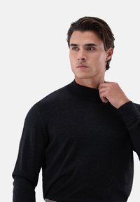 Junger Mann mit dunklen Haaren, der einen schwarzen Langarmpullover trägt, seinen Kragen ajustiert und nachdenklich zur Seite schaut, vor einem hellen Hintergrund.