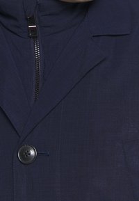 Giacca blu navy con colletto strutturato, chiusura con zip, un bottone e un tessuto texturizzato. Caratterizzata da una tasca sul petto e cuciture pulite.
