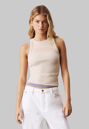 Top sin mangas de canalé beige con el logo de Calvin Klein, que presenta tirantes de un suave tono púrpura contrastante. Combinado con jeans de mezclilla blancos de cintura alta.