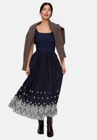 Maxikleid mit einem marineblauen Rock mit weißer Blumenstickerei, kombiniert mit einem taillierten marineblauen Oberteil und einer braunen Strickjacke.