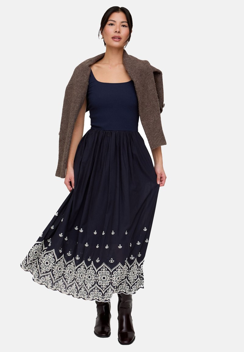 Maxikleid mit einem marineblauen Rock mit weißer Blumenstickerei, kombiniert mit einem taillierten marineblauen Oberteil und einer braunen Strickjacke.