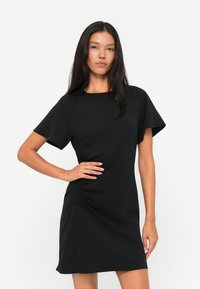 Schwarzes kurzärmliges Kleid aus einem glatten Stoff, mit seitlichem Raffen und einem leichten Schlitz am Saum für zusätzliche Details.