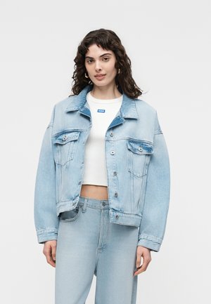 Femme aux cheveux bouclés bruns portant une veste en jean bleu clair surdimensionnée, un jean taille haute assorti et un T-shirt blanc court avec un logo bleu.