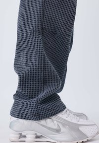 Pantalon gris foncé texturé avec un motif gaufré, associé à des chaussures Nike blanches avec un swoosh argenté et une semelle élastique.