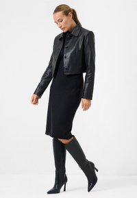 Zwarte leren cropped jack met knopen aan de voorkant, gedragen over een aansluitende zwarte jurk. Kniehoge zwarte stilettoboten maken de outfit compleet.