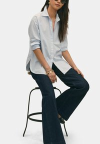 Gestreepte blauwe blouse met opgerolde mouwen, gecombineerd met donkere flared jeans. De persoon zit op een kruk, met zonnebril en sieraden.