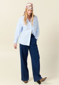Camicia a righe blu chiaro con bottoni abbinata a pantaloni in denim scuri larghi e stivali con tacco marroni; donna che indossa un foulard a fantasia.