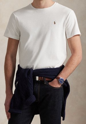 Man draagt een wit T-shirt met korte mouwen met een klein geborduurd logo, marineblauwe trui geknoopt om de taille, donkere spijkerbroek en een bruin leren horloge met een blauwe wijzerplaat.