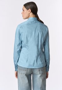 Camicia azzurro chiaro a maniche lunghe con un design attillato, polsini con bottoni e ricami sottili sul colletto. Tessuto morbido, in cotone.
