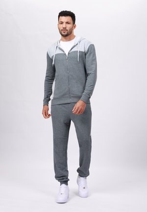 Grijze zip-up hoodie met een lichtgrijze yoke, gecombineerd met bijpassende broek. De stof is zachte fleece, met ribgebreide manchetten en witte sneakers.