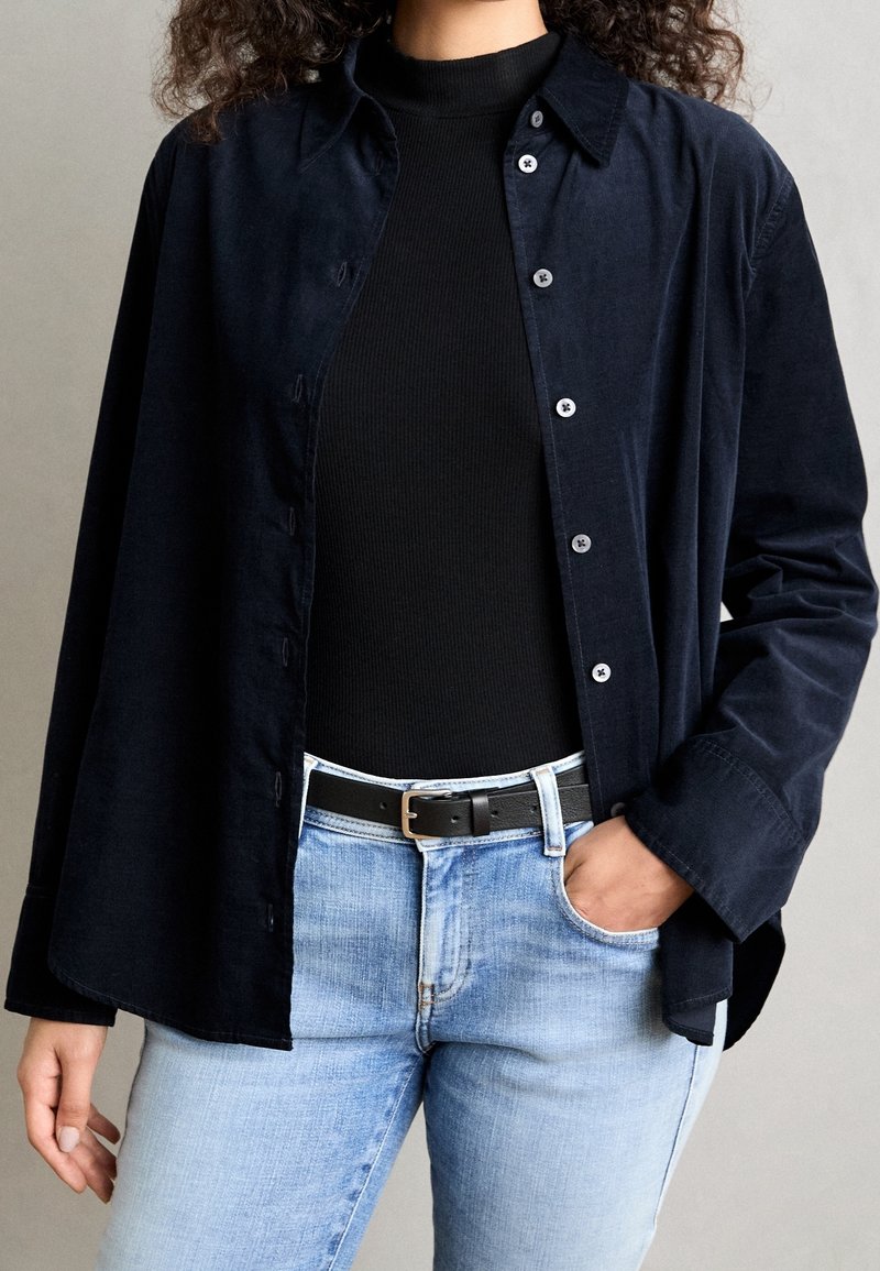 Personne portant une chemise noire à manches longues sous une veste bleu foncé déboutonnée, un jean bleu clair et une ceinture noire avec une boucle argentée, main dans la poche.