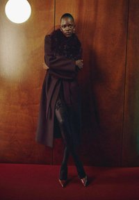 Cappotto lungo scuro con collare di pelliccia, collant neri con motivo a pizzo e tacchi a punta. Lo sfondo presenta pannelli di legno e una luce rotonda.