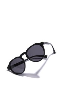 Čierne slnečné okuliare s hexagonálnymi šošovkami a šedým odtieňom. Rám s matným povrchom a úzke nožičky. Nápis "Vyrobené v Španielsku" je viditeľný vo vnútri.