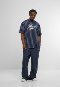 Vīrietis, kurš valkā tumši zilu svītrainu t-kreklu un atbilstošas brīvā stila bikses ar baltām kedām, stāv pret vienkāršu fonu.