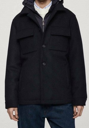 Homme portant une veste noire à capuche avec poches à rabat à l'avant, fermeture éclair et boutons, associée à un jean bleu foncé et un pull en maille blanc.