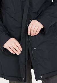 Sort parka med en tekstureret overflade, lynlås- og trykknaplukninger, der har sidelommer og fleeceforede manchetter. Hænder justerer åbningen.