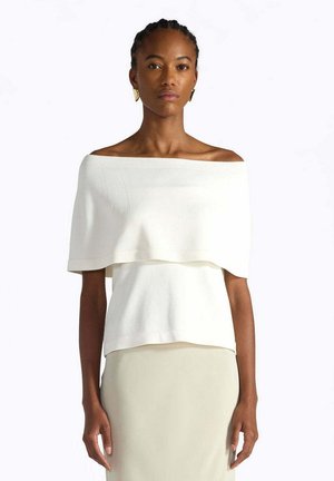 Donna con capelli intrecciati che indossa una maglietta bianca a strati con spalle scoperte e una gonna beige, in piedi davanti a uno sfondo chiaro e uniforme.