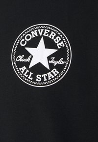 Černá tkanina s bílým kruhovým logem se hvězdou, s nápisy "CONVERSE ALL STAR" a "Chuck Taylor" v stylizovaném písmu.