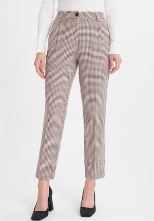 Femme portant un pantalon taille haute à carreaux beige, noir et rouge, associé à un pull blanc côtelé et des chaussures noires à talons hauts.