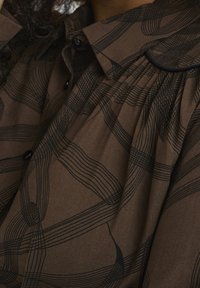 Brown blouse with black wave-like pattern, featuring a collar and gathered detailing. Soft fabric with button closures.