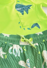 Tricou de înot verde neon cu design de rechin și buzunar. Pantaloni scurți verzi din material texturat, cu modele de caracatiță și pește, accentuați cu șnur de ajustare.