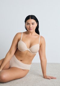 Beige naadloze bh met dunne verstelbare bandjes en een subtiele sluiting aan de voorkant, gedragen met hoge beige broekjes op een lichte tapijtvloer.