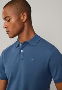 Modrá polo košile s klasickým límečkem, na hrudi malá vyšitá modrá logo a knoflíkovou aplikací. Bavlněná látka s hladkým povrchem.
