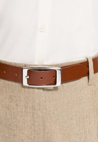 Cintura in pelle marrone con fibbia rettangolare in argento indossata sopra pantaloni beige con texture e una camicia bianca a pulsanti infilata dentro.