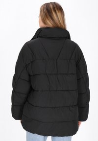 Kvinde med langt lysebrunt hår iført en sort polstret jakke og lyseblå jeans, vist bagfra mod en hvid baggrund.