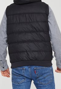 Gilet d'avorio nero con design trapuntato orizzontale, abbinato a una maglietta a maniche lunghe grigia e jeans blu. Presenta un orlo a costine e un marchio discreto.