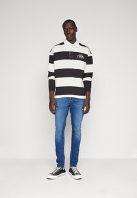 Polo de manga larga a rayas blancas y negras con cuello blanco, combinado con jeans skinny azules y zapatillas negras. Logo en la camiseta visible.