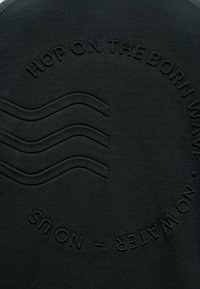 Embossovaná čierna tkanina s tromi vlnitými čiarami a okruhovým textom, ktorý znie "NASCITE NA BORN WAVE • NO WATER – NO US."