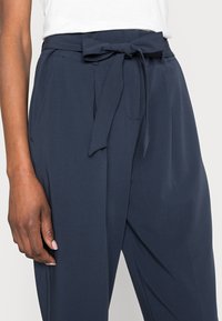 Calças ajustadas azul-marinho com uma textura suave, apresentando um cinto laço na cintura, bolsos laterais e um corte descontraído.