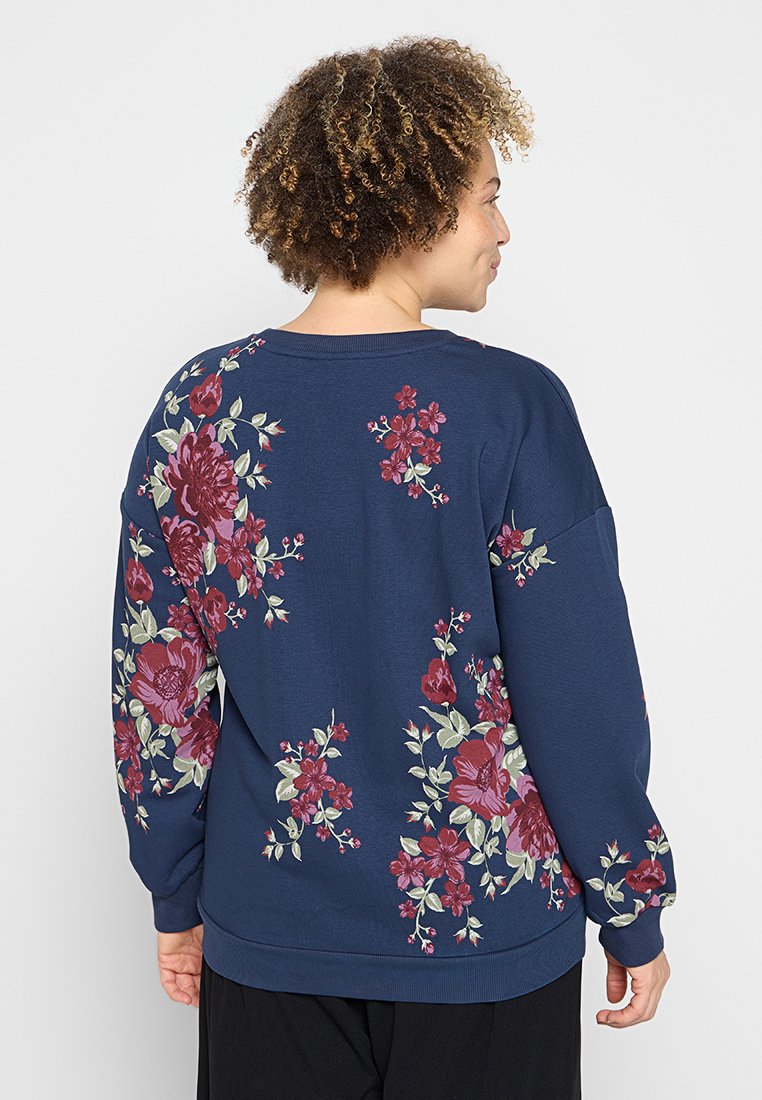 Vrouw met krullend haar, gekleed in een marineblauwe sweatshirt versierd met rode en roze bloempatronen, gezien van achteren tegen een effen achtergrond.