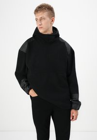 Felpa nera con cappuccio realizzata in morbido fleece, con eleganti dettagli in pelle nera sui gomiti e sulle spalle; presenta una vestibilità oversize e un colletto alto.